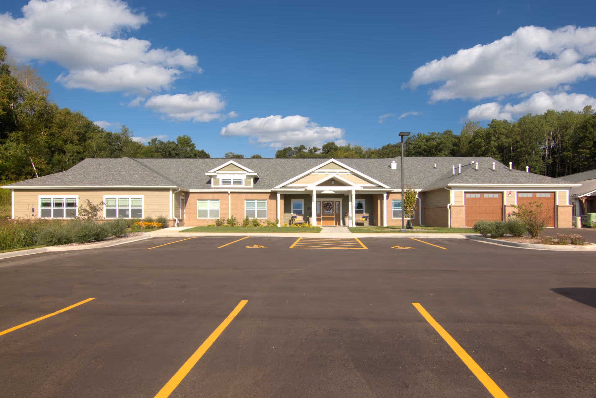 Front exterior view of a single-story memory care facility building with a gray roof, beige siding, and brick accents. The entrance has a covered porch with white columns and two benches. There is a large parking lot in front with marked handicap parking spaces. Trees and greenery surround the building under a partly cloudy blue sky.