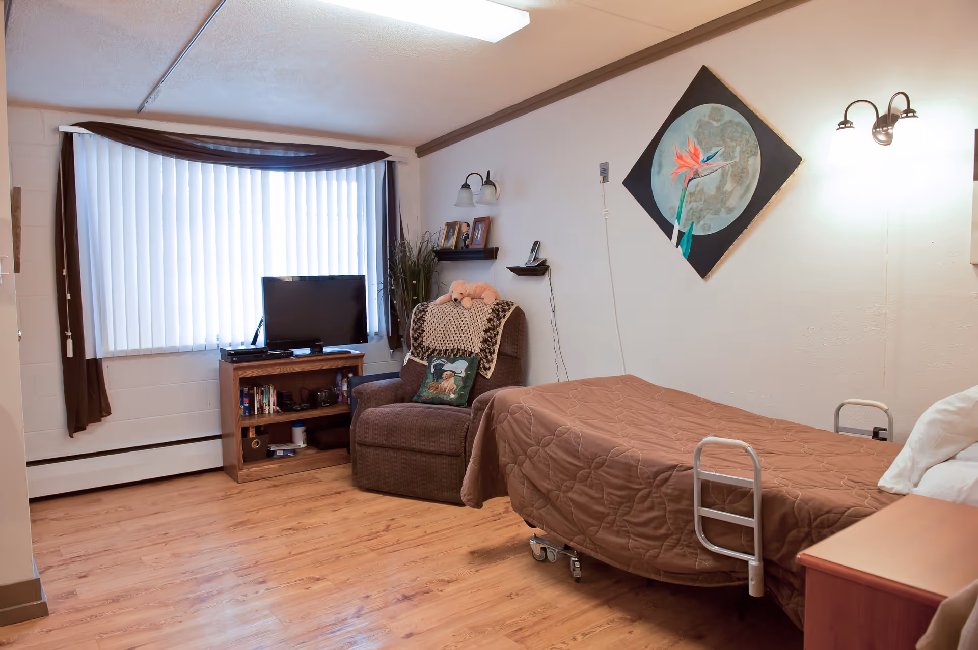 A cozy senior living bedroom with a single bed covered in a brown quilt, a comfortable armchair with a decorative pillow and a blanket, a small wooden TV stand with a flat-screen television, books, and other items. The room has a large window with vertical blinds and brown curtains, light wood flooring, and a wall painting of a bird of paradise flower.