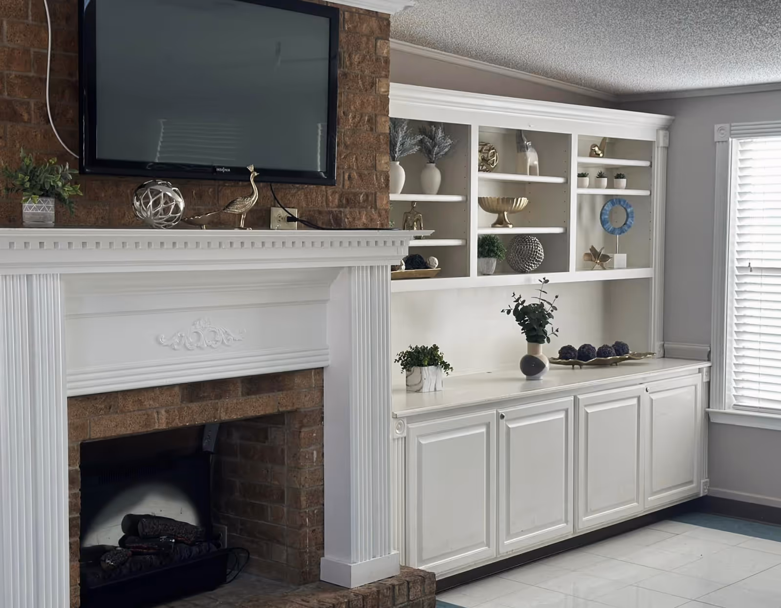 A cozy living room area featuring a brick fireplace with a white mantel. Above the mantel is a mounted flat-screen TV. To the right of the fireplace is a built-in white cabinet with shelves displaying decorative items such as vases, plants, and sculptures. A window with white blinds is visible on the far right, allowing natural light into the room.