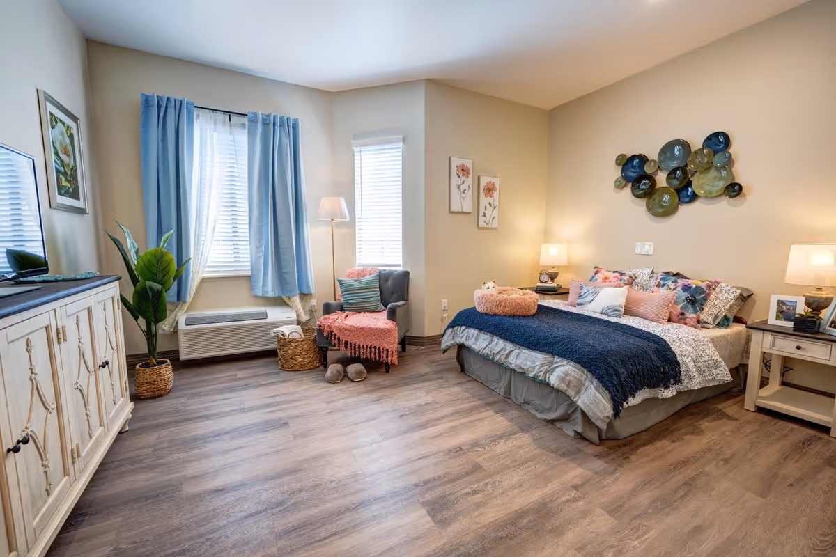 A cozy bedroom in a senior living facility featuring a neatly made bed with multiple pillows and a dark blue throw blanket. The room has light-colored walls with decorative wall art above the bed. There is a nightstand with a lamp and framed photo beside the bed. A comfortable armchair with a pink throw blanket and slippers sits near two windows with blue curtains. A large cabinet with a TV on top is positioned opposite the bed. The floor is wood-style laminate, and a potted plant adds greenery near the windows.