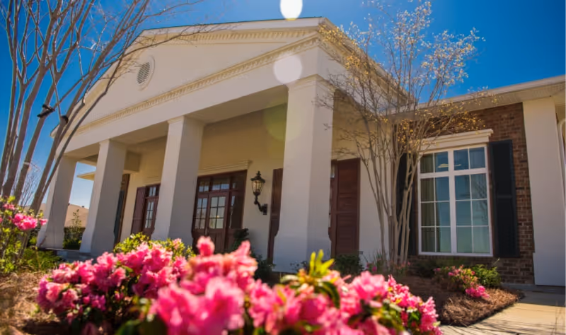 Front entrance of a rehabilitation center with white columns, large windows, and pink flowers in the foreground.
