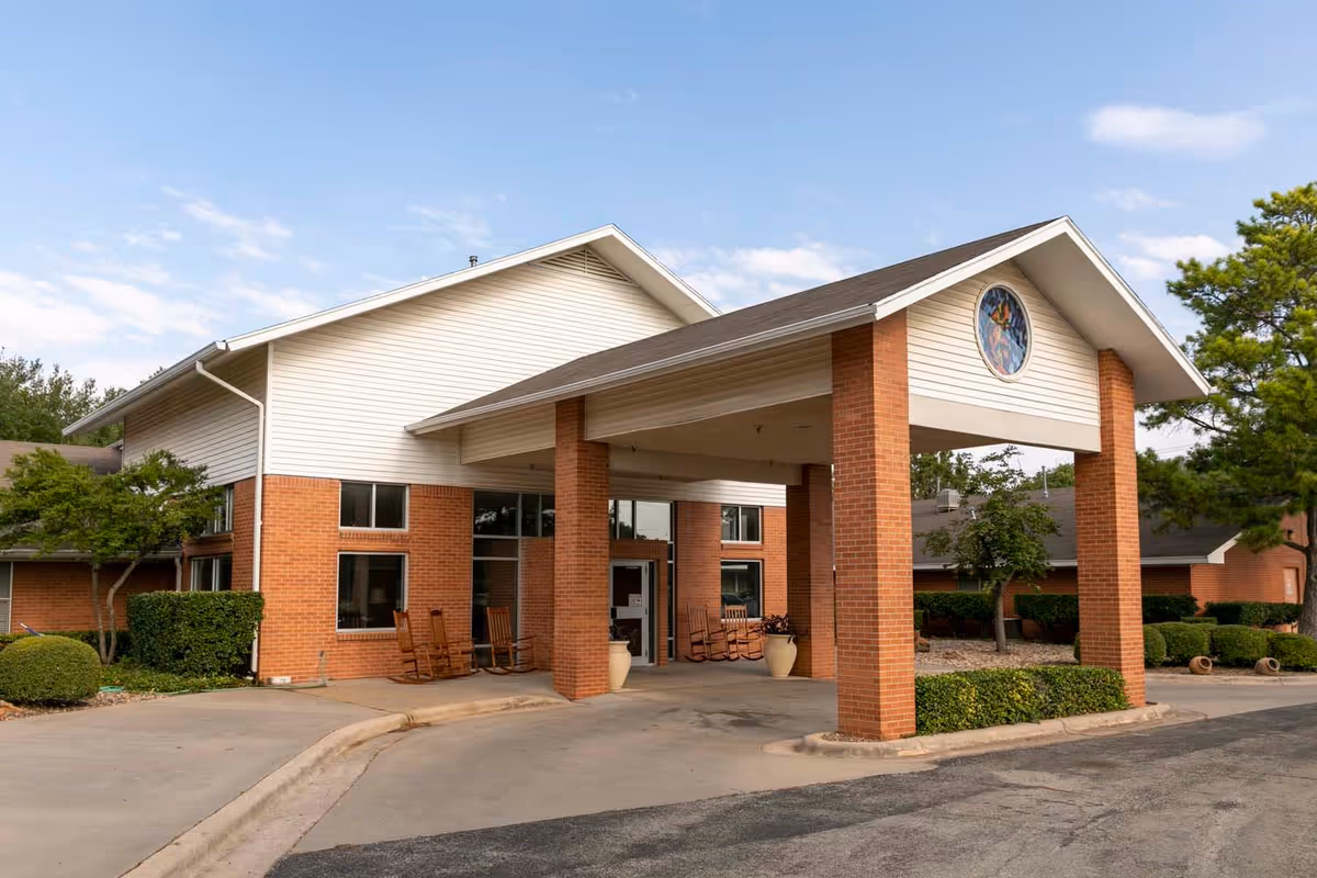 Exterior view of Wisteria Place Assisted Living facility showing a covered entrance with brick pillars, rocking chairs, and well-maintained landscaping under a partly cloudy sky.