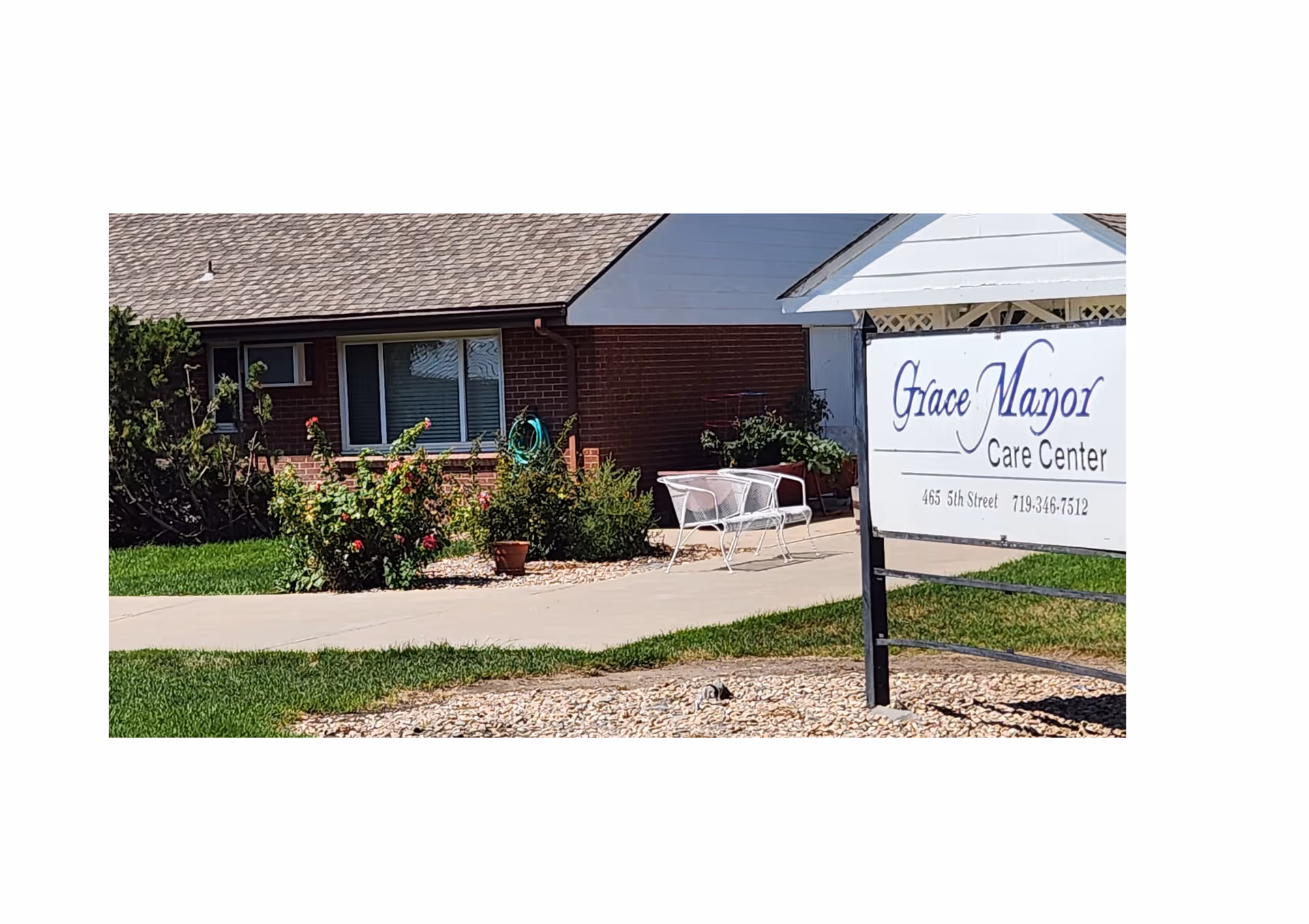 Exterior view of Grace Manor Care Center showing a brick building with a window, a garden with flowers and shrubs, a white bench on a concrete pathway, and a sign with the facility's name, address, and phone number.