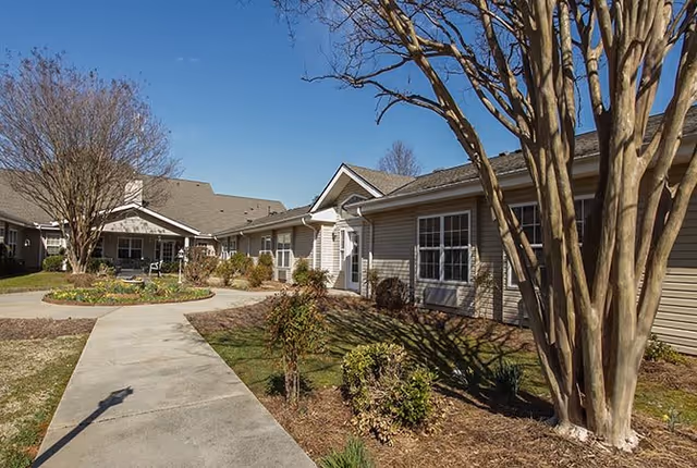 Exterior view of a single-story senior living facility building with beige siding, multiple windows, and a concrete walkway leading through landscaped grounds with trees and shrubs under a clear blue sky.