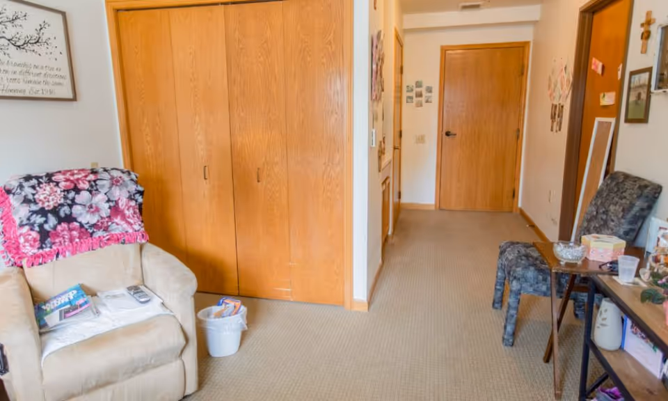 Interior living area of an assisted living suite with a beige recliner covered by a floral throw, wooden closet doors, and a hallway leading to a closed door.