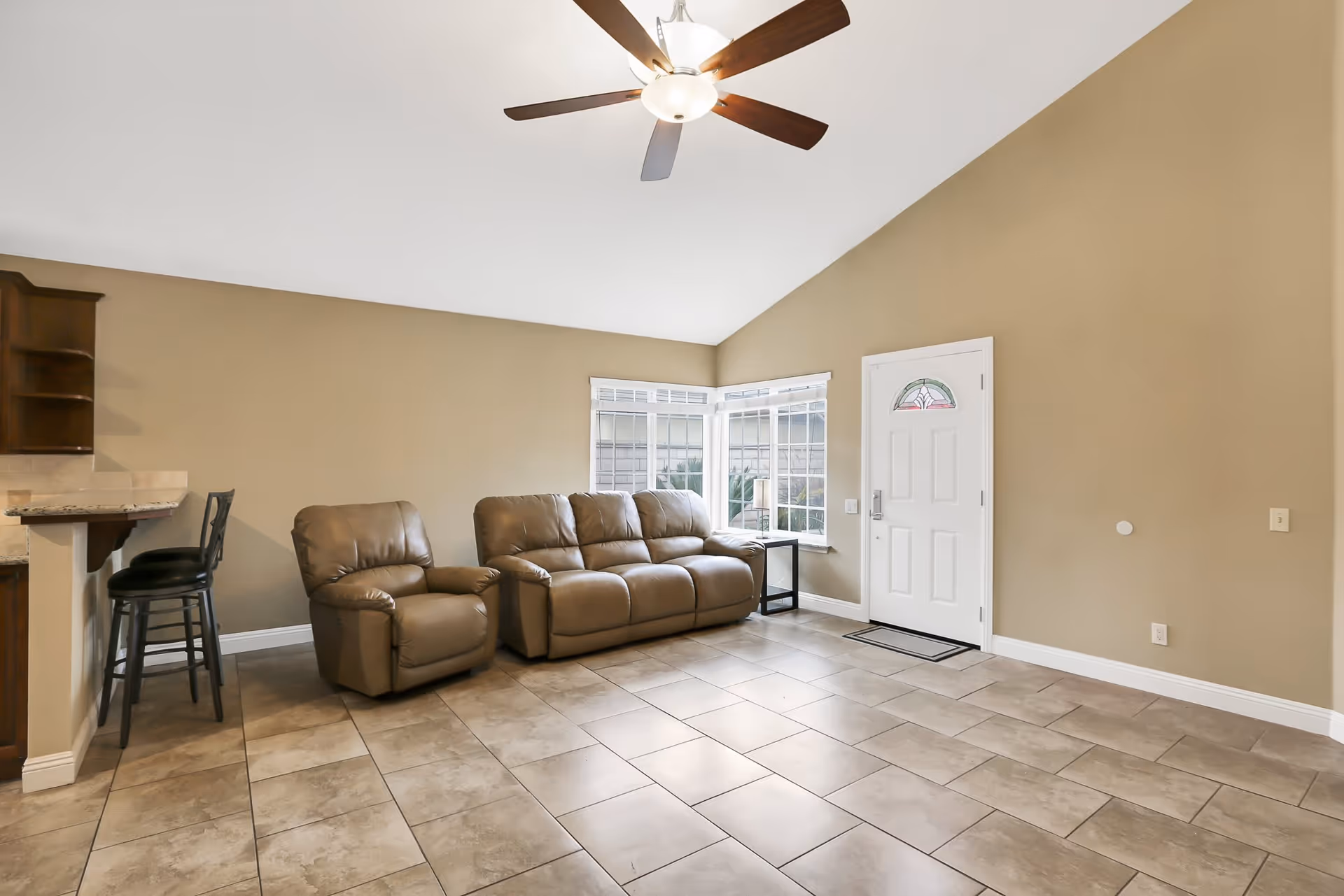 A living room with beige tiled floor, tan leather sofa and armchair, a ceiling fan with light, a white front door with a decorative glass window, and a corner window with white blinds. There is a small black side table with a lamp next to the sofa and two black bar stools at a granite countertop on the left side.