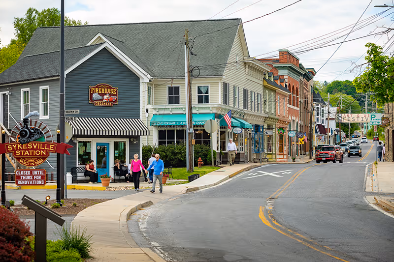 A small town street scene with a few people walking on the sidewalk near a building with a sign that reads 'Firehouse Creamery'. The street curves to the right and is lined with various shops and buildings. A sign in the foreground reads 'Sykesville Station, Built in 1868, Closed Until Thurs for Renovations'. Trees and greenery are visible along the street.
