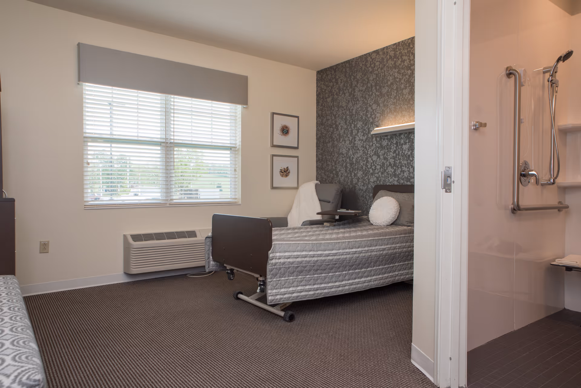 A senior living facility bedroom with a single bed covered in a gray quilt, a gray armchair with a white throw blanket, two framed pictures on the wall, a large window with blinds, and an adjacent accessible bathroom with grab bars and a handheld showerhead.