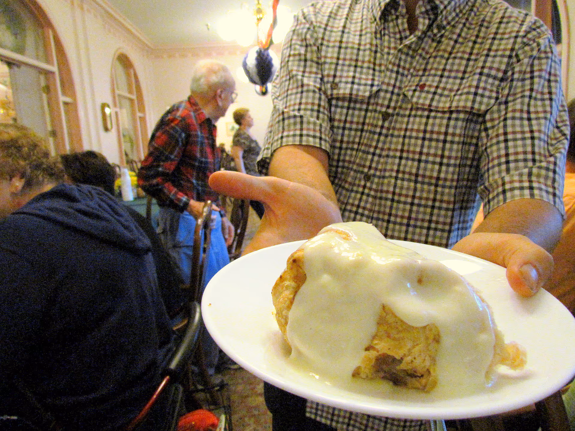 A person presents a plate with a pastry covered in white sauce while several people sit and stand in a crowded dining room.
