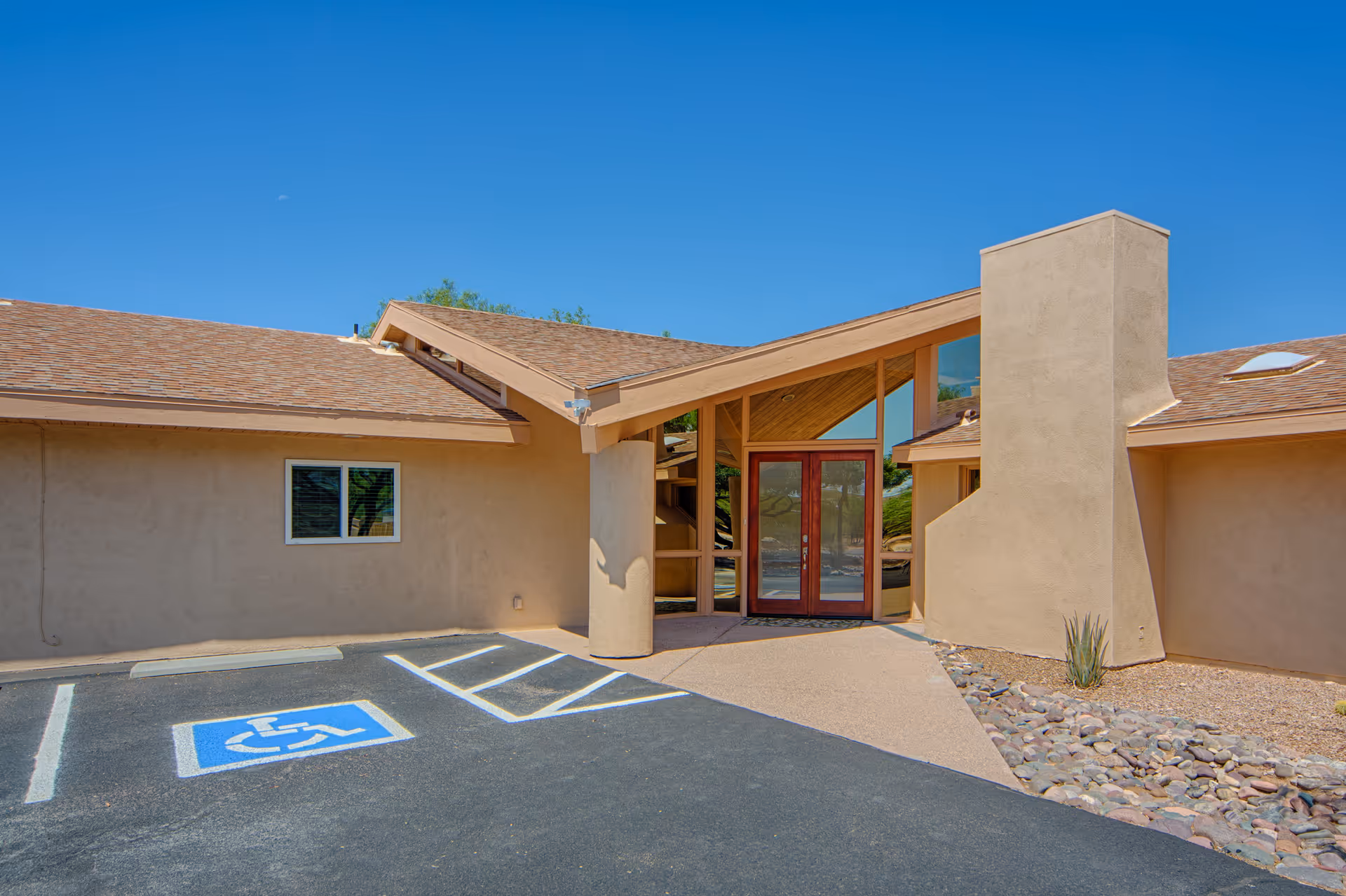 Exterior view of The Gardens at San Carlos facility entrance with a clear blue sky, a handicapped parking space in front, beige stucco walls, a sloped roof, large glass double doors, and a stone-lined landscaping area.