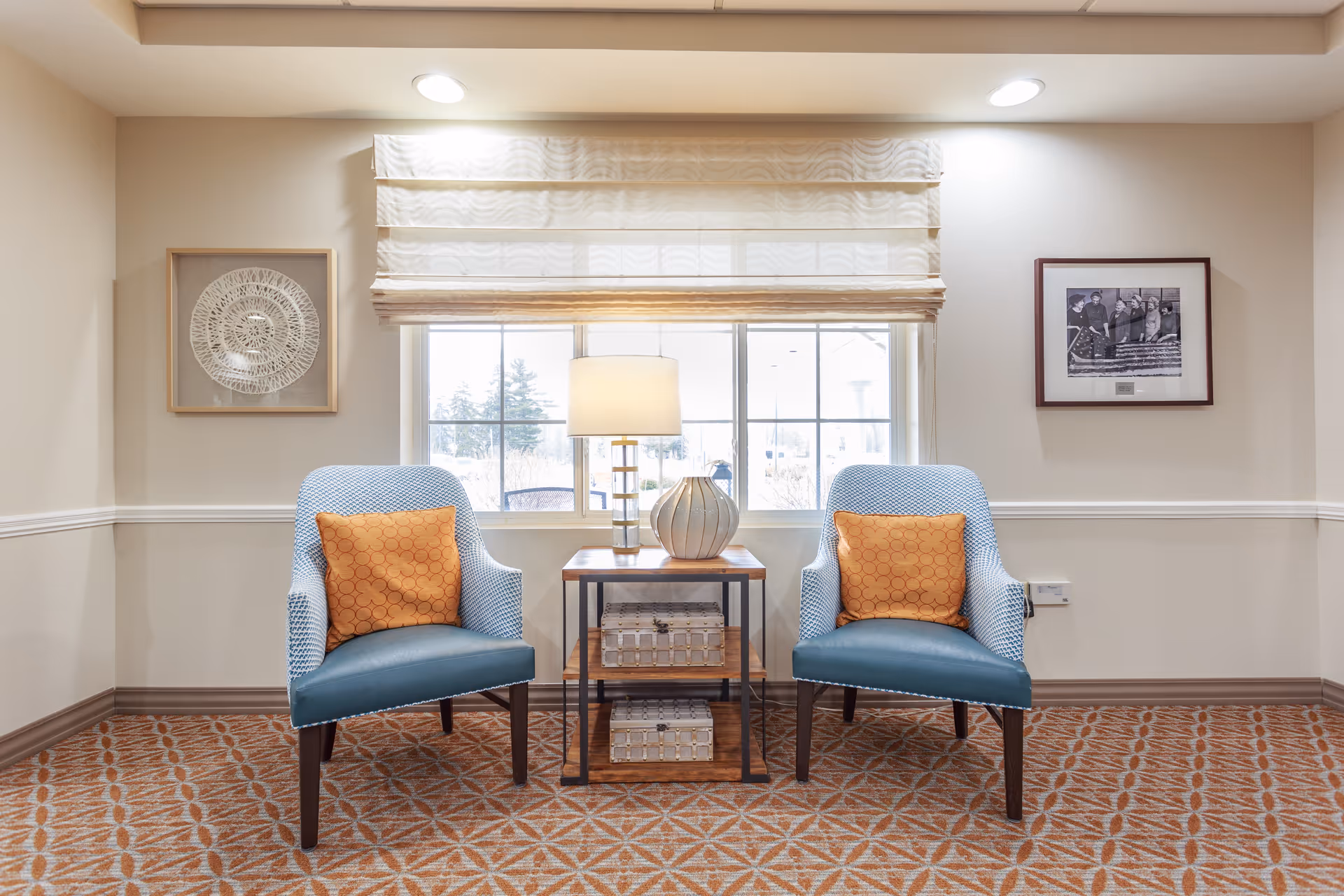 A cozy seating area with two blue armchairs, each with an orange patterned pillow, positioned on either side of a wooden side table. The table holds a decorative lamp and a vase, with two woven baskets on the lower shelves. Behind the seating area is a window with a white Roman shade, and the walls are adorned with framed artwork. The floor is covered with a patterned carpet.