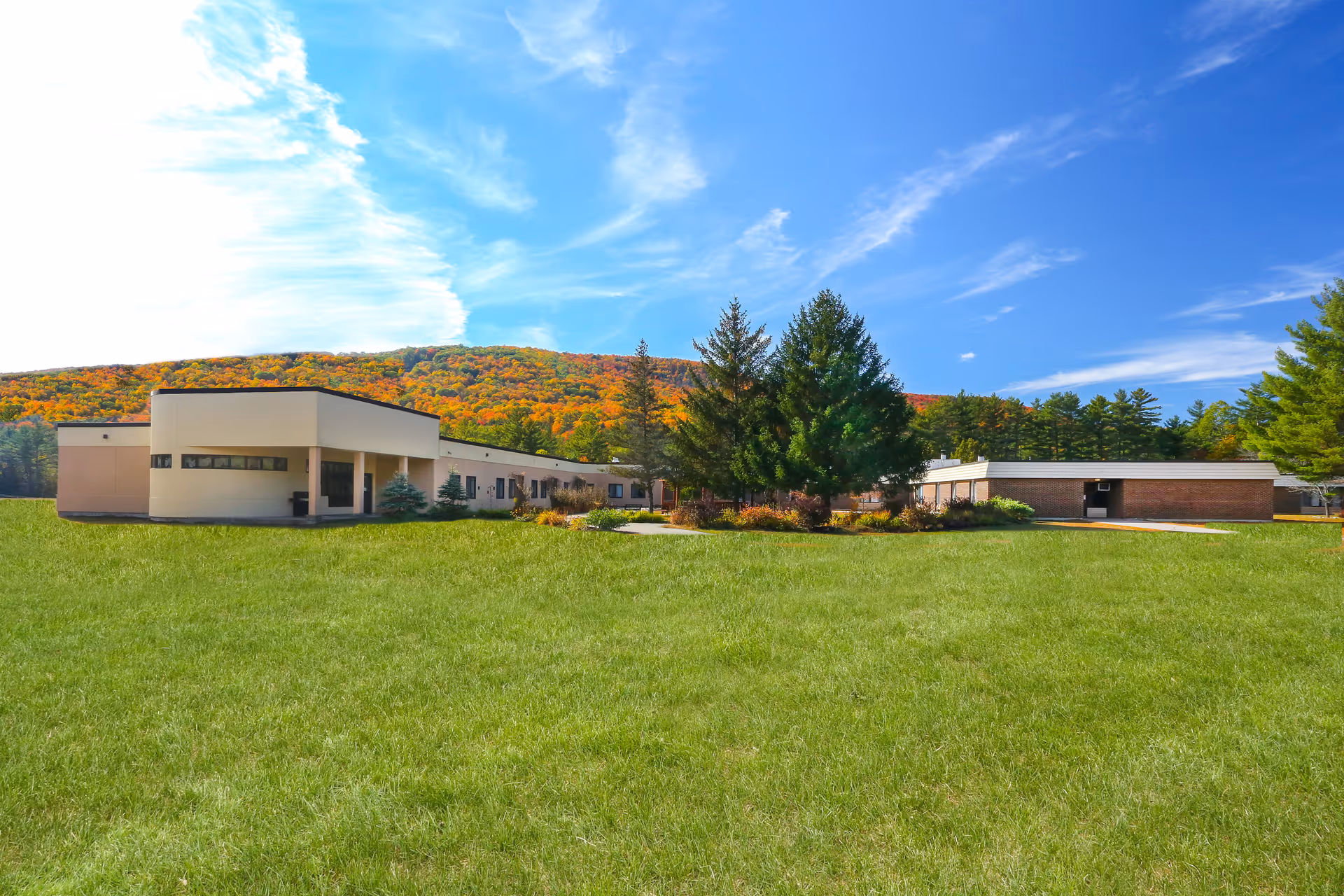 Single-story senior living facility building with a wide grassy lawn, evergreen trees, and wooded hills under a blue sky.
