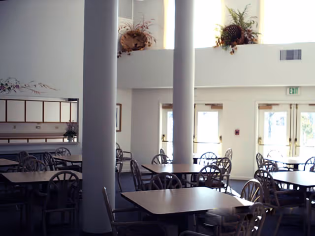A dining area with multiple square tables and chairs arranged around them. The room has large white pillars, high ceilings, and large windows letting in natural light. There are decorative plants placed on a ledge near the ceiling and an exit door in the background.
