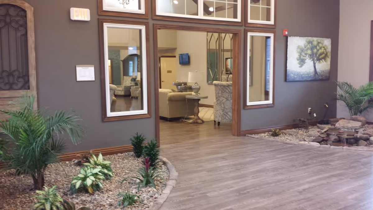 Indoor area of a senior living facility with a small indoor garden featuring various plants and rocks. The room has wood flooring and gray walls with large windows and a doorway leading to a lounge area with beige and patterned armchairs, a side table with a lamp, and a wall-mounted TV. There is a painting of a tree on the wall and an exit sign above a door.