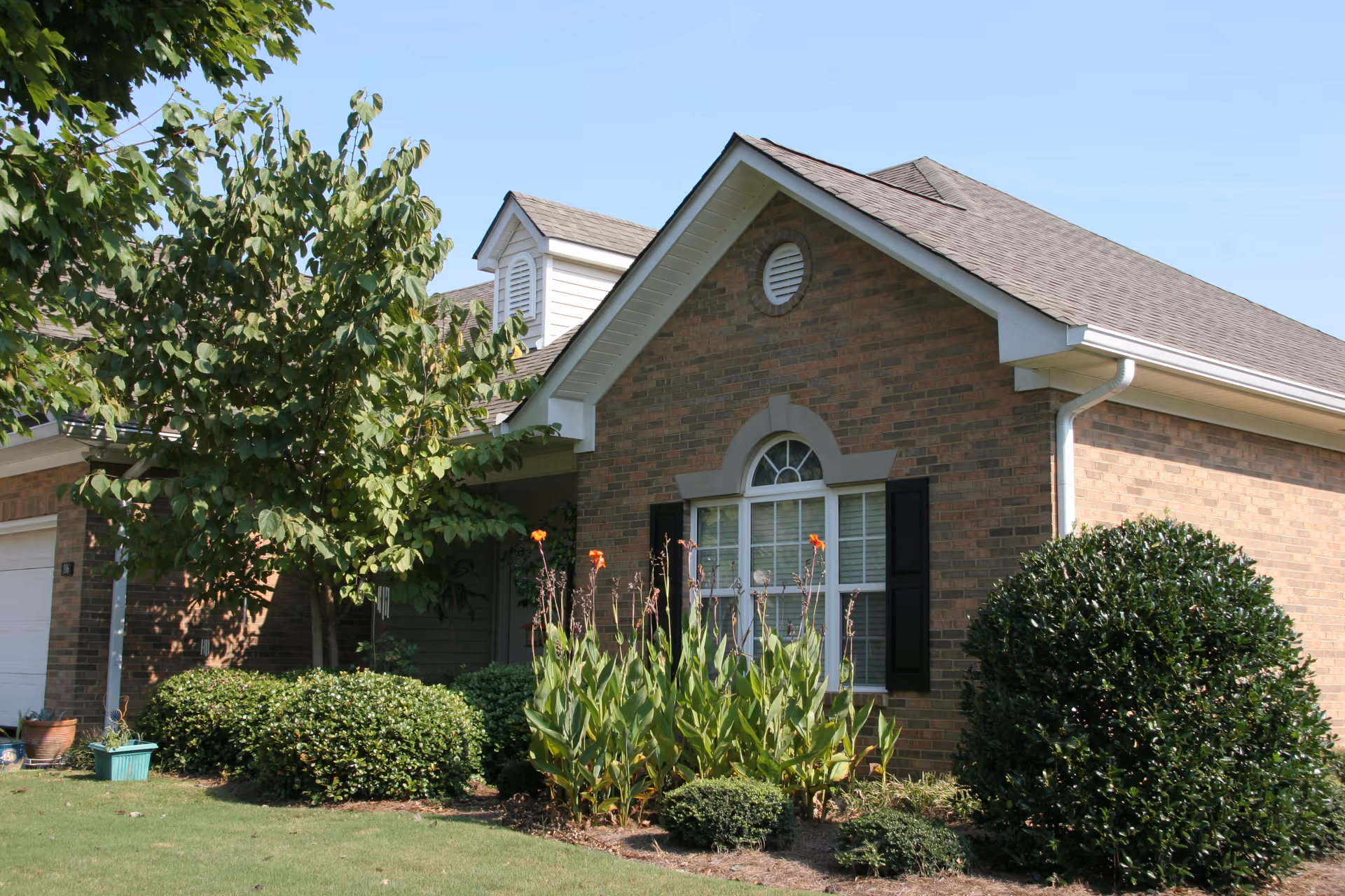 Exterior view of a single-story brick house with a gable roof, a large window with black shutters, and a well-maintained front yard featuring green shrubs, flowering plants, and a tree under a clear blue sky.