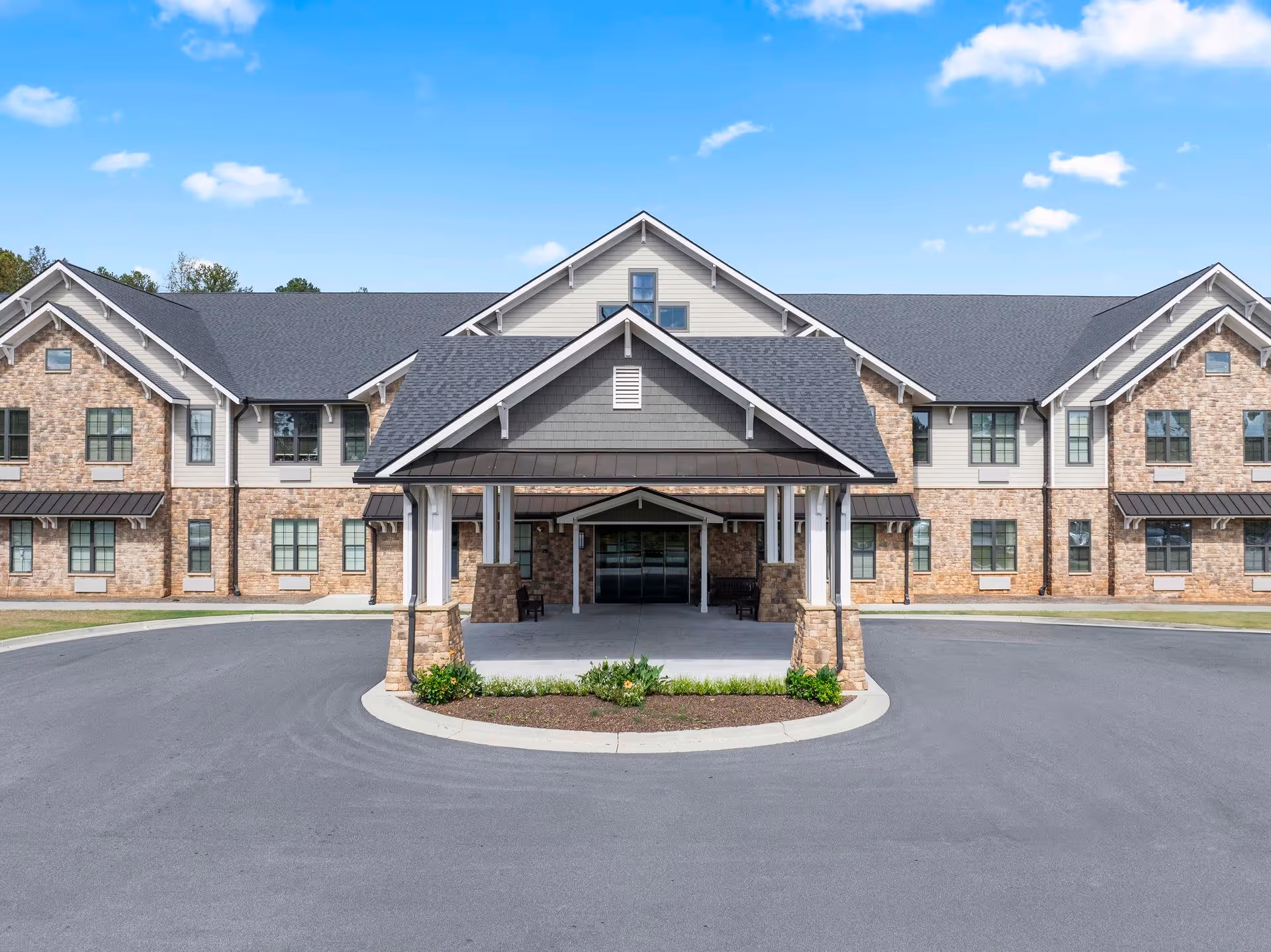 Front exterior view of a two-story senior living facility building with a covered entrance, stone and siding facade, multiple windows, and a circular driveway under a blue sky with scattered clouds.