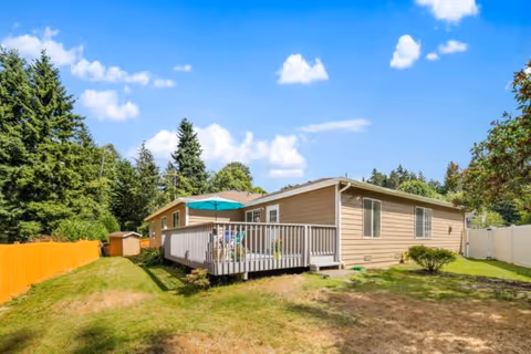 Single-story beige house with a wooden deck featuring a green umbrella and outdoor chairs, surrounded by a grassy yard with trees and a wooden fence under a blue sky with scattered clouds.