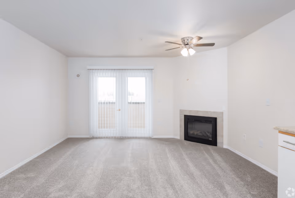 Empty living room with beige carpet, white walls, a ceiling fan with lights, a small fireplace with tile surround, and glass doors with vertical blinds leading to an outdoor area.