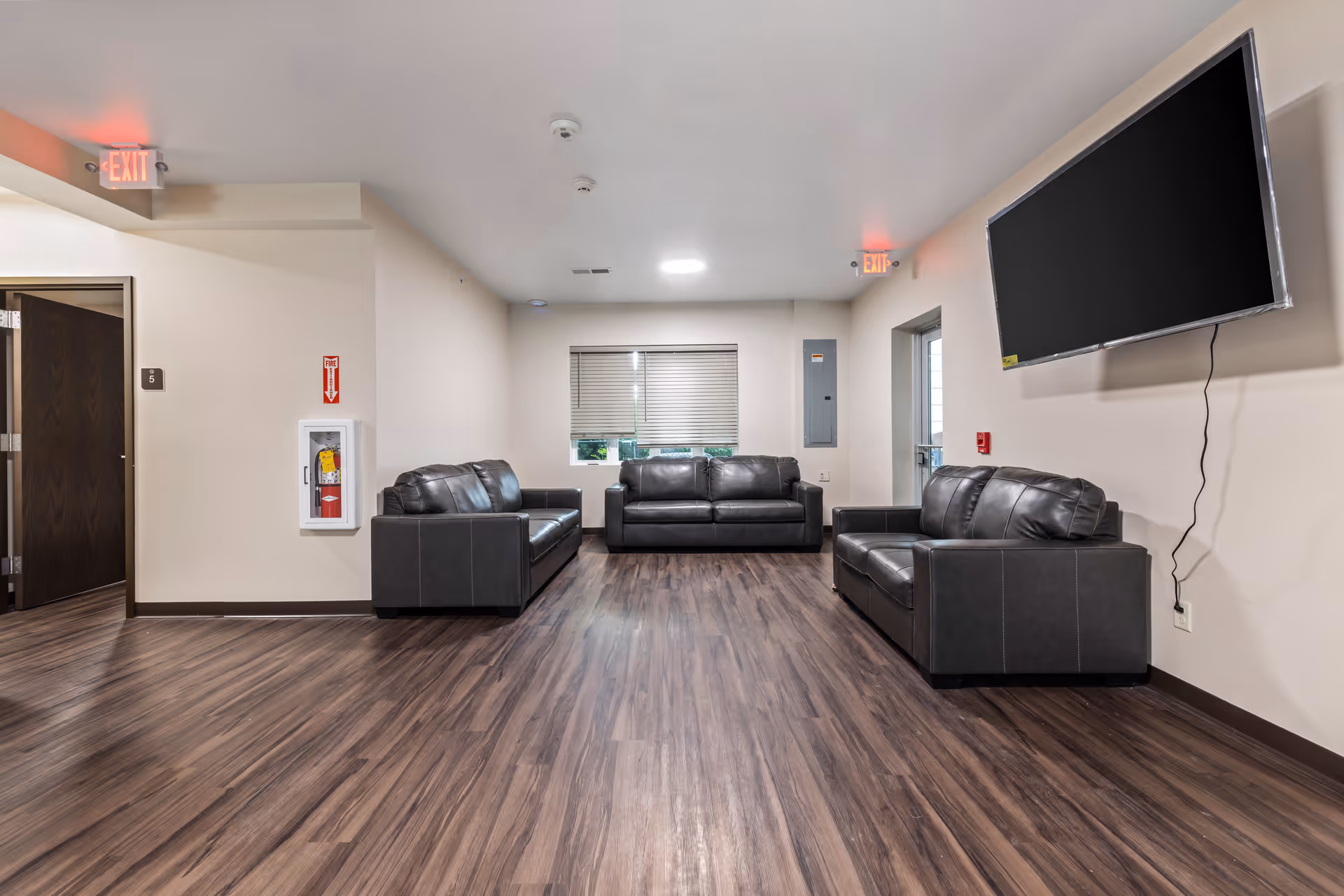 A spacious living room area with three black leather sofas arranged around the room. The floor has dark wood laminate, and the walls are painted light beige. A large flat-screen TV is mounted on the right wall with visible cables hanging down. There are two exit signs illuminated in red above doors on the left and right sides. A window with closed blinds is centered on the far wall, and a fire extinguisher is mounted on the left wall.