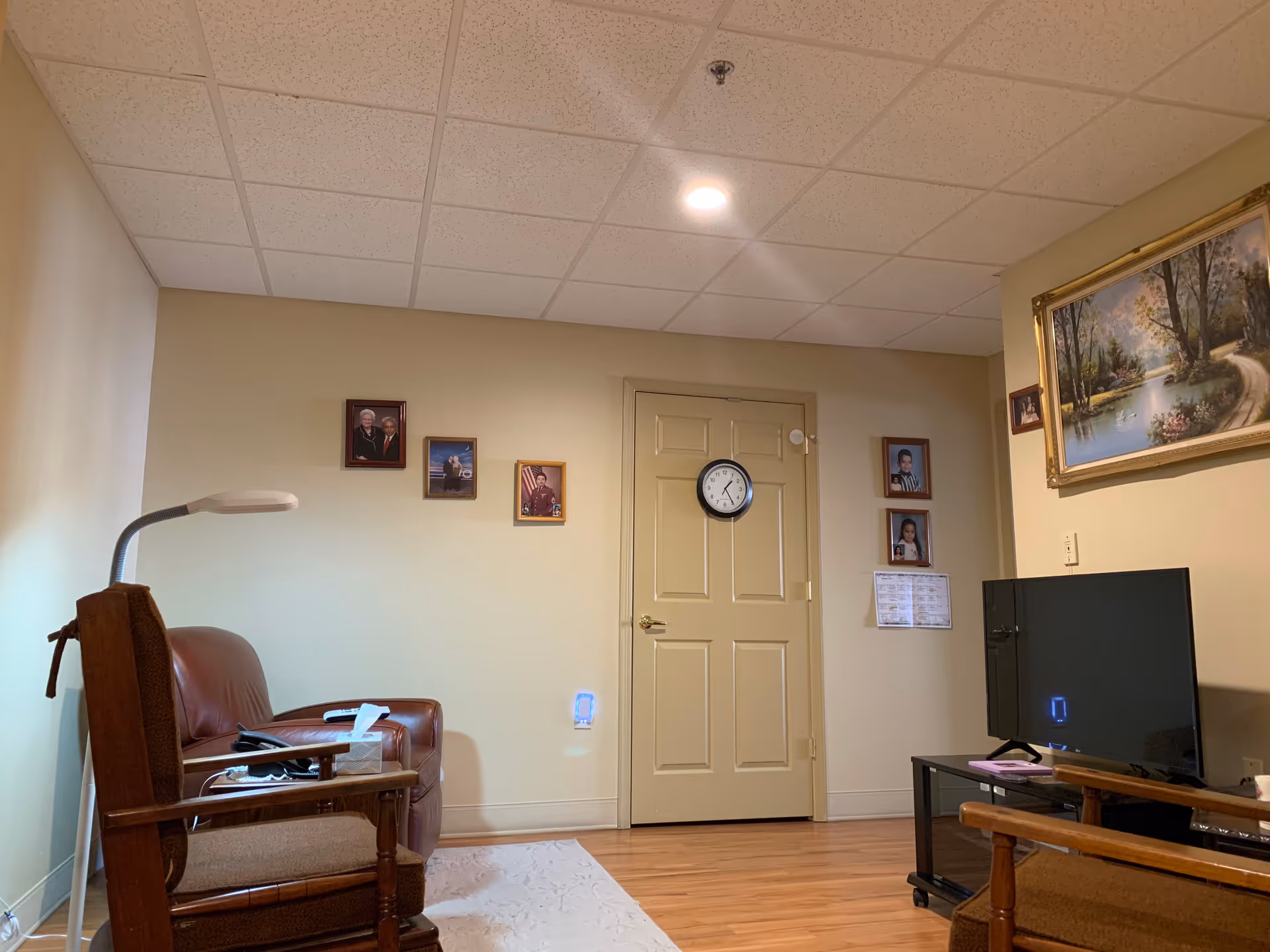 A cozy living room with beige walls and wooden flooring. There are two chairs, one with a brown cushion and the other a leather recliner. A floor lamp is positioned next to the chairs. On the wall, there are several framed photographs and a clock hanging on a closed door. A flat-screen TV sits on a black stand beneath a framed landscape painting.