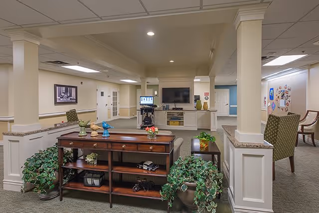 A spacious senior living common area with beige walls and carpeted floors. The room features a wooden console table with decorative items and plants on and around it. In the background, there is a large flat-screen TV mounted on a white entertainment unit, a computer workstation, and several chairs arranged along the walls. The ceiling has recessed lighting and a drop ceiling design.
