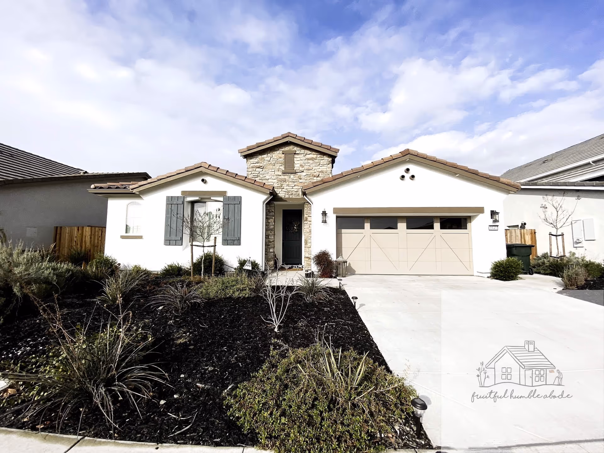 Front exterior of a single-story house with a two-car garage, stone entry tower, and landscaped front yard under a partly cloudy sky.