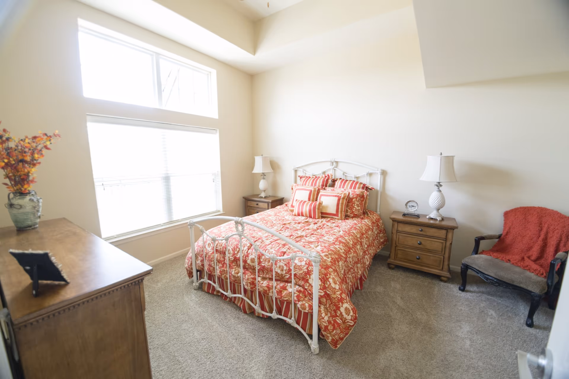 A bright bedroom with a white metal bed frame and a red floral and striped bedspread. The bed has multiple matching pillows. There are two wooden nightstands on either side of the bed, each with a white lamp. A wooden dresser with a vase of flowers and a framed photo is on the left side of the room. A cushioned chair with a red throw blanket is positioned near the right nightstand. The room has beige walls, carpeted floor, and large windows letting in natural light.