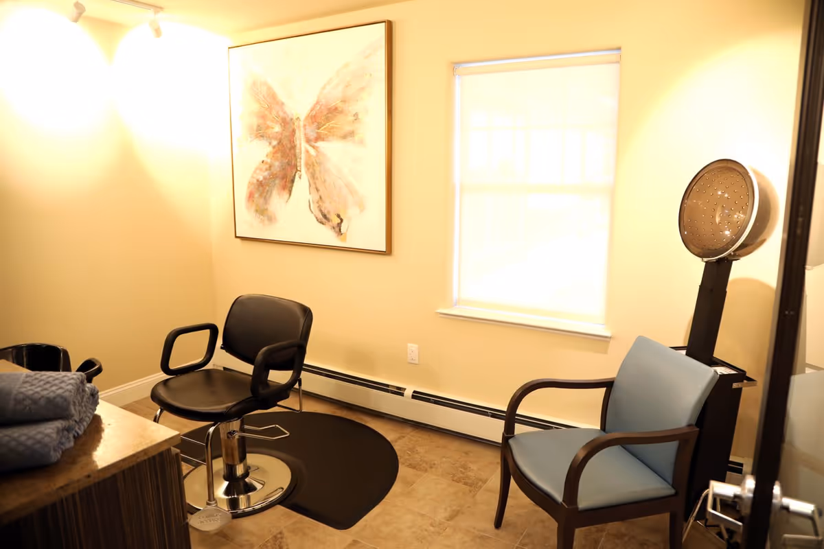 A small salon room with a black salon chair on a black mat, a blue cushioned chair, a hair dryer hood, a window with a white blind, a countertop with folded blue towels, and a large framed butterfly artwork on the wall.