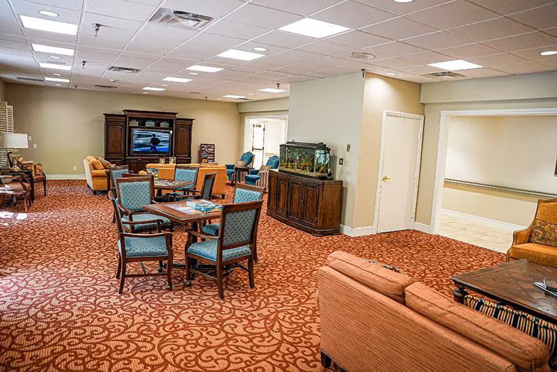 A spacious senior living common area with patterned red carpet, multiple seating arrangements including blue cushioned chairs around a table, orange sofas, and a large wooden entertainment center with a television. There is a fish tank on a wooden cabinet against a wall, and the room is well-lit with ceiling lights.