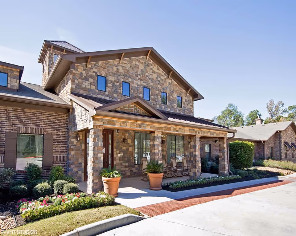 Exterior view of a two-story building with stone and brick facade, featuring a covered porch with rocking chairs and potted plants. The building is surrounded by landscaped greenery and flowers under a clear blue sky.