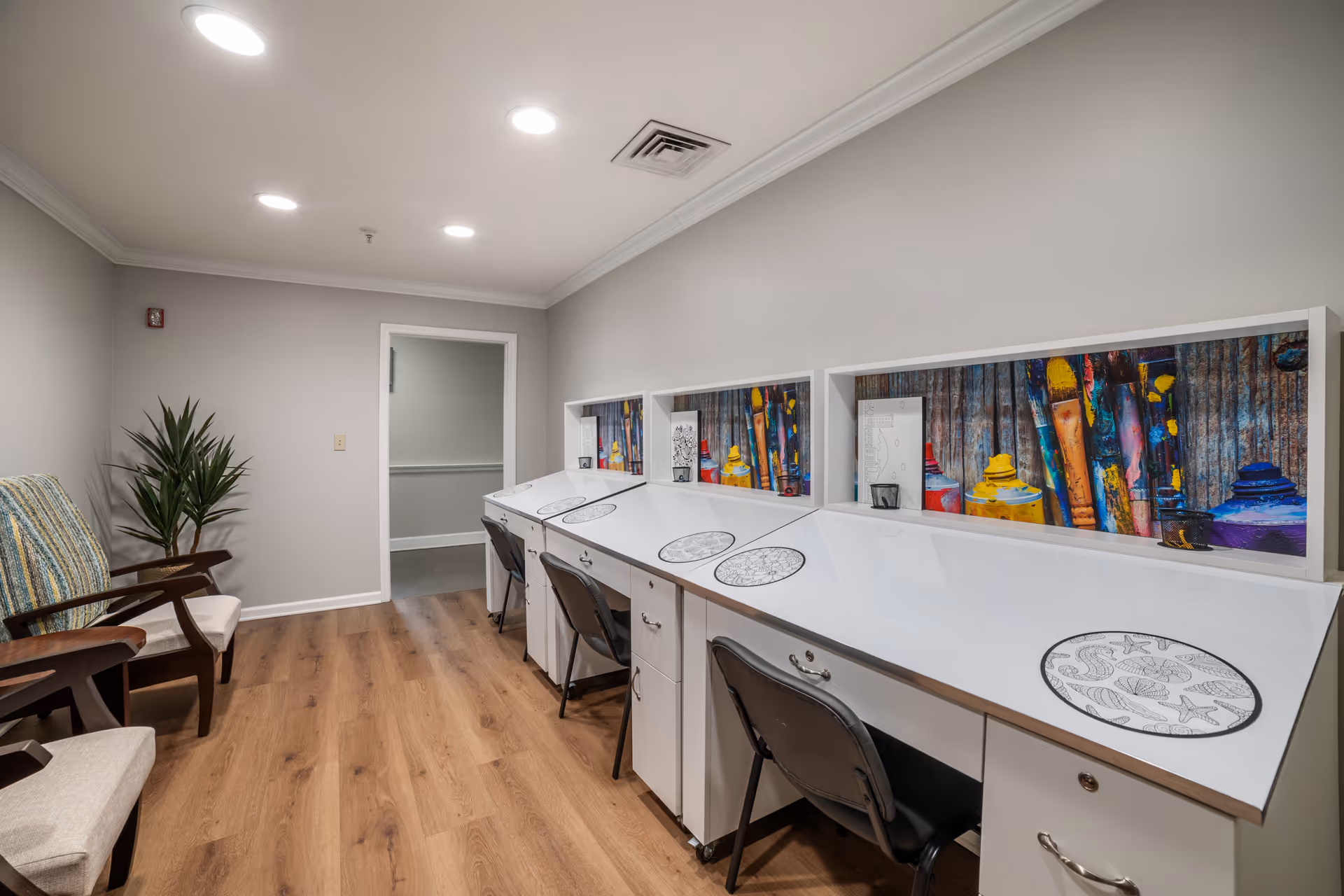 A well-lit room with a long white angled desk featuring four workstations, each with a black chair and circular coloring sheets. Behind the desk are colorful art-themed panels with paintbrushes and paint tubes. On the left side of the room are two cushioned chairs with wooden armrests and a tall green plant. The floor is wooden, and the walls are painted light gray with white trim.