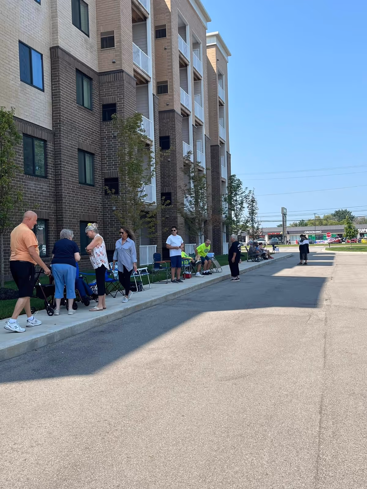 People gathered along the sidewalk outside a multi-story brick apartment building with balconies under a clear blue sky.