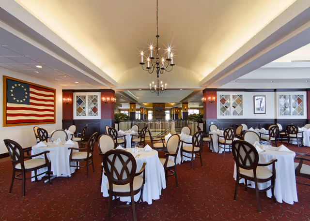 A spacious dining room with multiple round tables covered with white tablecloths and set with glassware and napkins. The room features elegant wooden chairs with cushioned seats, a red carpet with a subtle pattern, and a high ceiling with two chandeliers. On the left wall, there is a framed Betsy Ross American flag, and the walls have decorative panels and framed artwork.