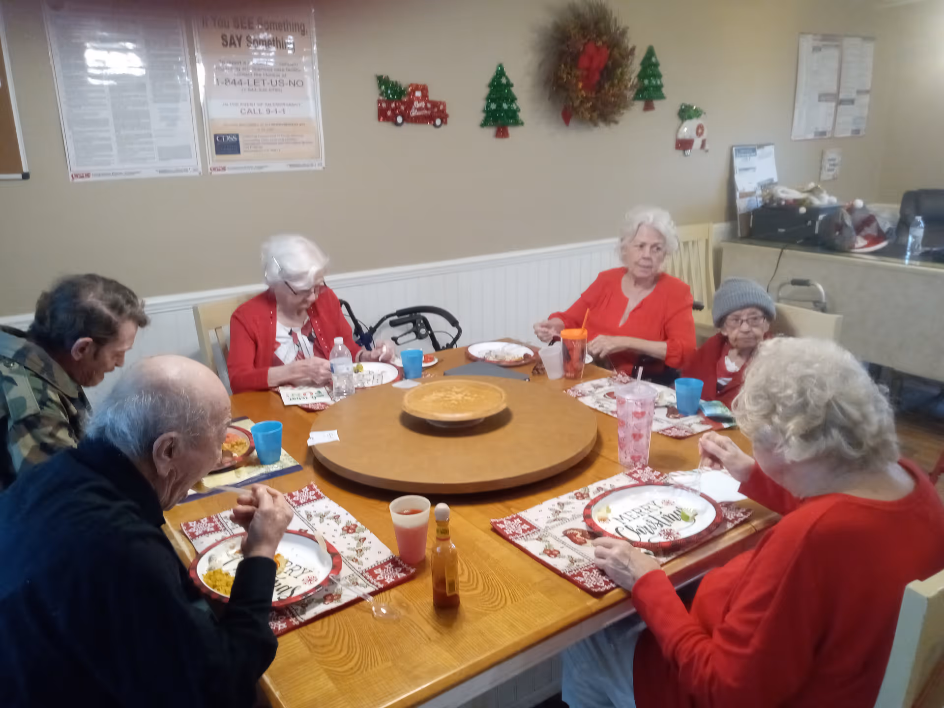 A group of elderly people sitting around a wooden dining table eating a meal together. The table has Christmas-themed placemats and plates, and there are holiday decorations on the wall including a wreath and small Christmas trees. The room has beige walls and some informational posters on the wall.