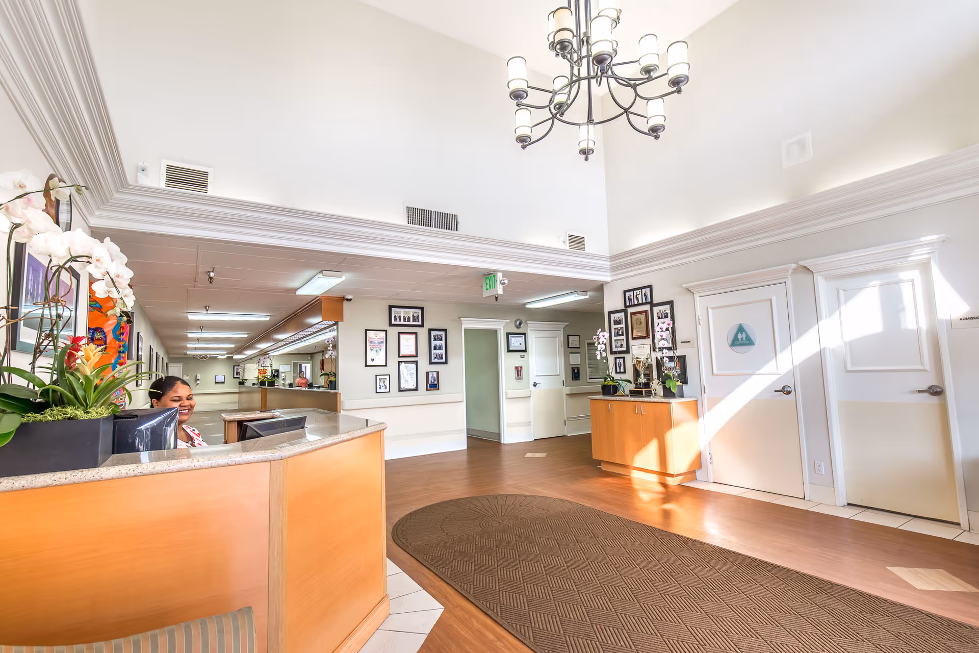 Reception area of Windsor Care Center of Cheviot Hills featuring a smiling receptionist behind a wooden desk with a granite countertop. The space has high ceilings with crown molding, a chandelier, framed pictures on the walls, and a brown patterned rug on the wooden floor. There are two white doors on the right side and a small cabinet with plants and flowers.