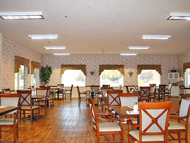 A spacious dining room with wooden floors and multiple wooden tables and chairs arranged neatly. The walls are decorated with floral wallpaper and windows with valance curtains allow natural light to brighten the room. There are some plants and decorative items along the walls.