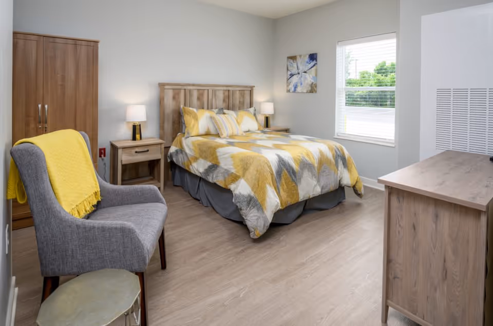 A bright bedroom featuring a wooden bed with yellow-and-gray bedding, matching nightstands, a gray armchair with a yellow throw, a wardrobe, and a window.