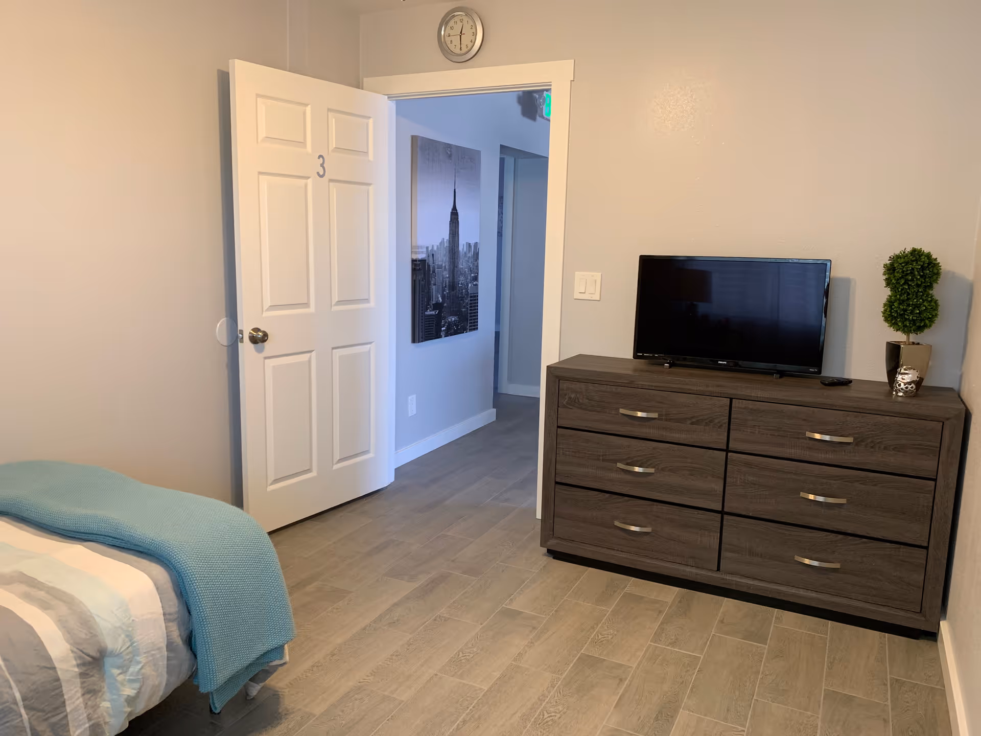 A simple bedroom with a bed partially visible on the left side covered with a striped blanket and a blue throw. There is a white door marked with the number 3 that is open, revealing a hallway with a black and white cityscape picture on the wall. On the right side of the room, there is a dark wooden dresser with six drawers, a flat-screen TV on top, a small plant, and a decorative owl figurine. A clock is mounted above the door on the wall.