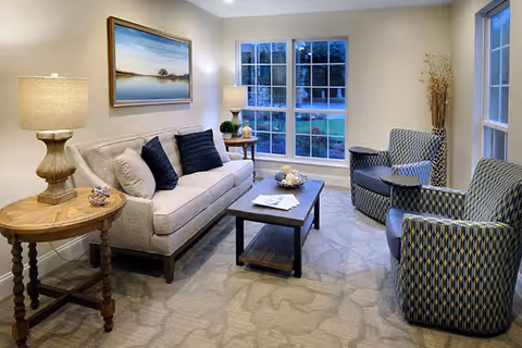 A cozy living room with a beige sofa adorned with dark and light cushions, two patterned armchairs, a wooden coffee table with decorative items, two side tables each with a lamp, a large window showing an outdoor view, and a landscape painting on the wall.