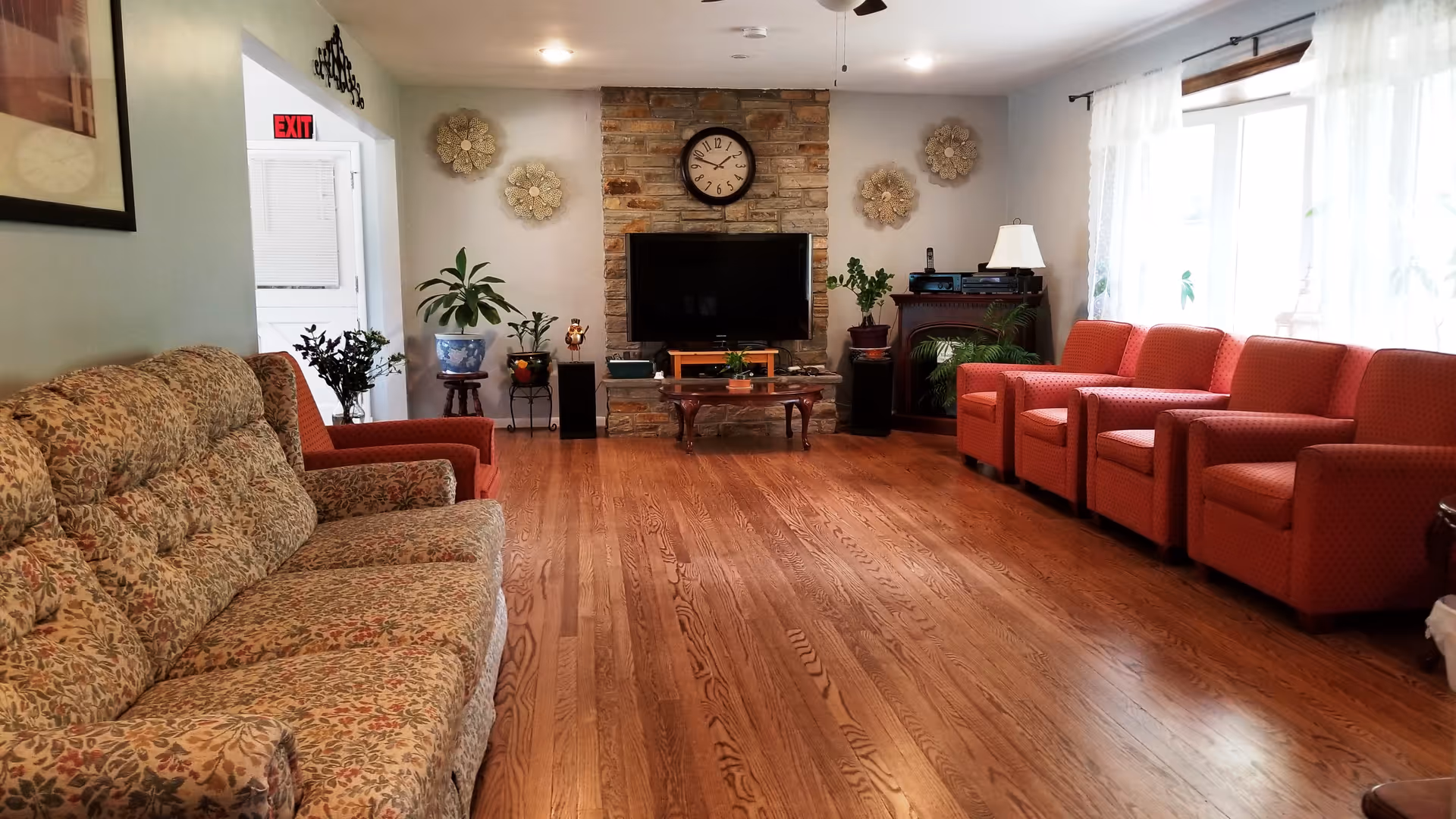 A spacious living room with wooden flooring, featuring a floral patterned sofa on the left and a row of five red armchairs on the right near large windows with white curtains. A stone fireplace with a clock above it and a flat-screen TV mounted on the wall is centered at the far end of the room. Several potted plants and decorative wall hangings are also visible.