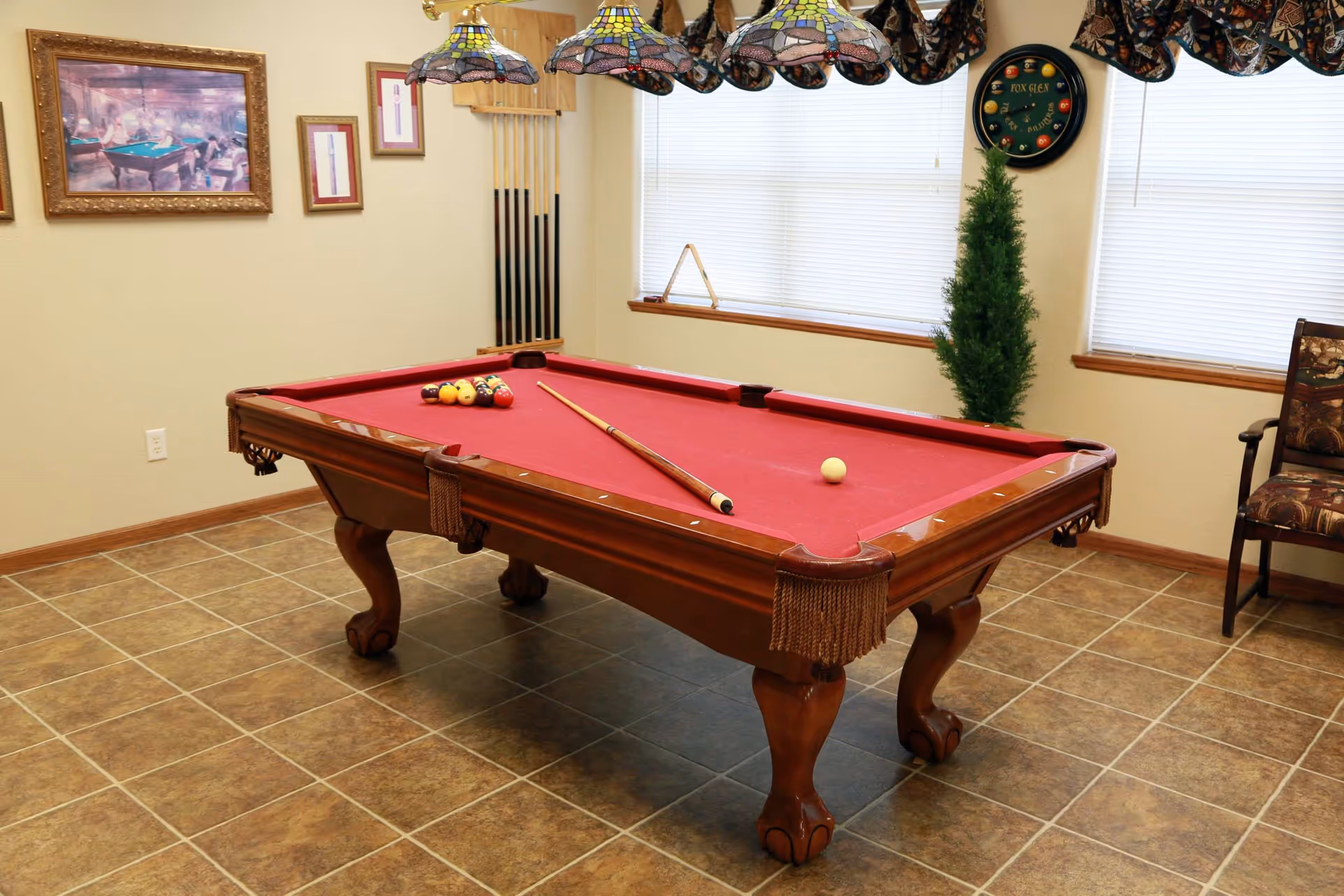 A red-felt pool table with cues and balls in a tiled game room with framed art, windows, and seating.