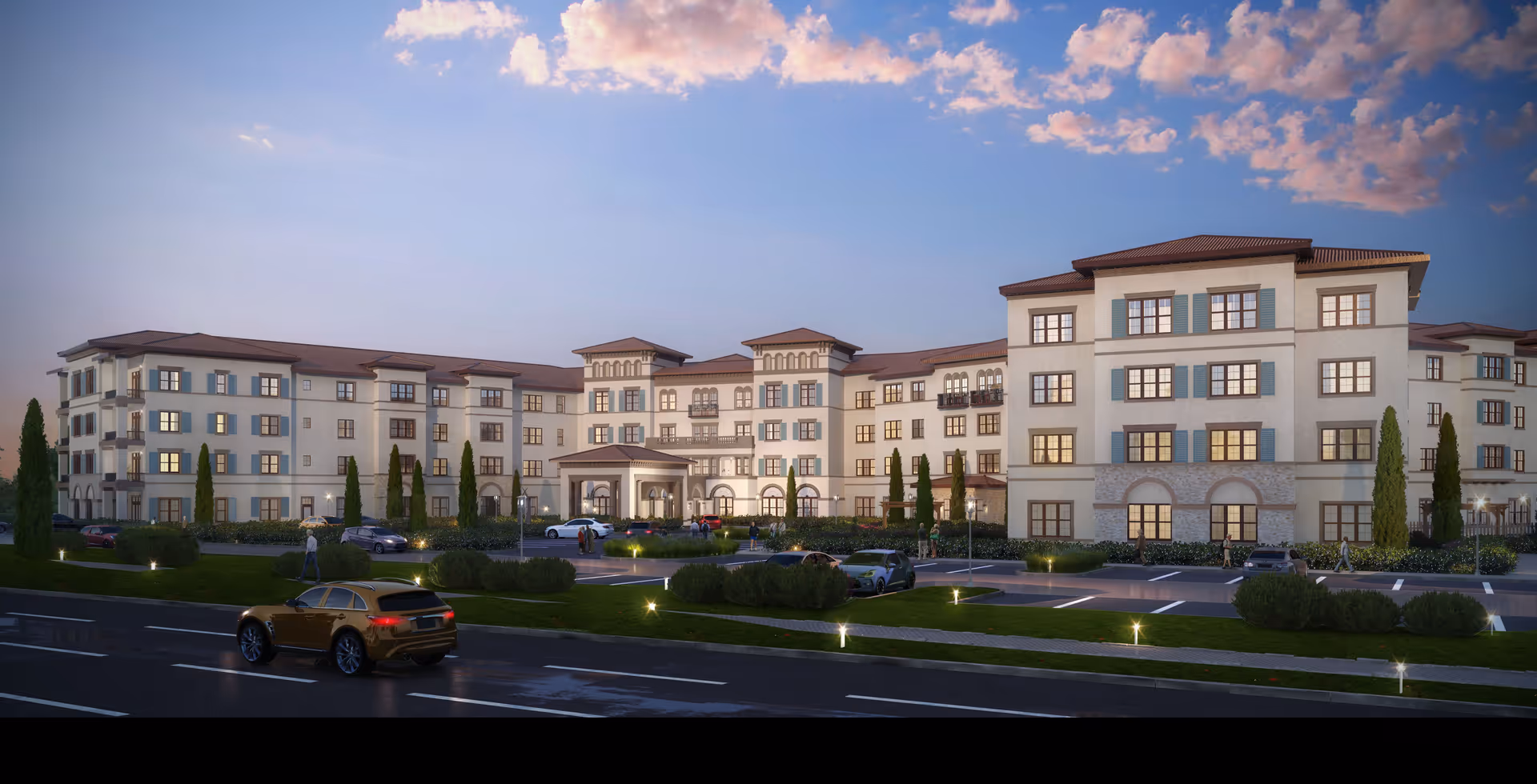 Exterior view of a large, multi-story senior living facility building with Mediterranean-style architecture, featuring beige walls, blue shutters, and a red-tiled roof. The building is surrounded by landscaped greenery, a parking lot with several cars, and a road in the foreground under a partly cloudy sky at dusk.