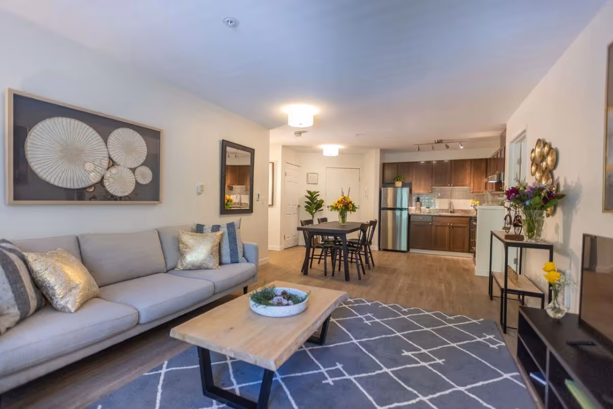 A modern living space featuring a light gray sofa with decorative pillows, a wooden coffee table on a patterned rug, a dining table with four chairs, and a kitchen area with dark wood cabinets and stainless steel appliances. The room is decorated with wall art, flowers, and plants, creating a cozy and inviting atmosphere.