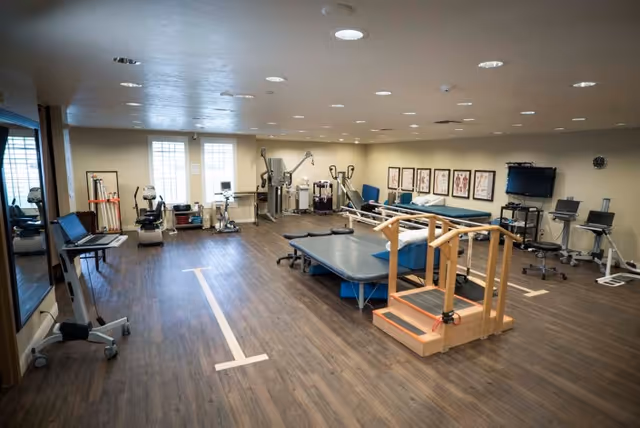 A spacious physical therapy or rehabilitation room with exercise equipment, therapy tables, parallel bars for walking practice, anatomical charts on the wall, and a television mounted on a stand. The room has wood flooring and is well-lit with ceiling lights and natural light from windows.