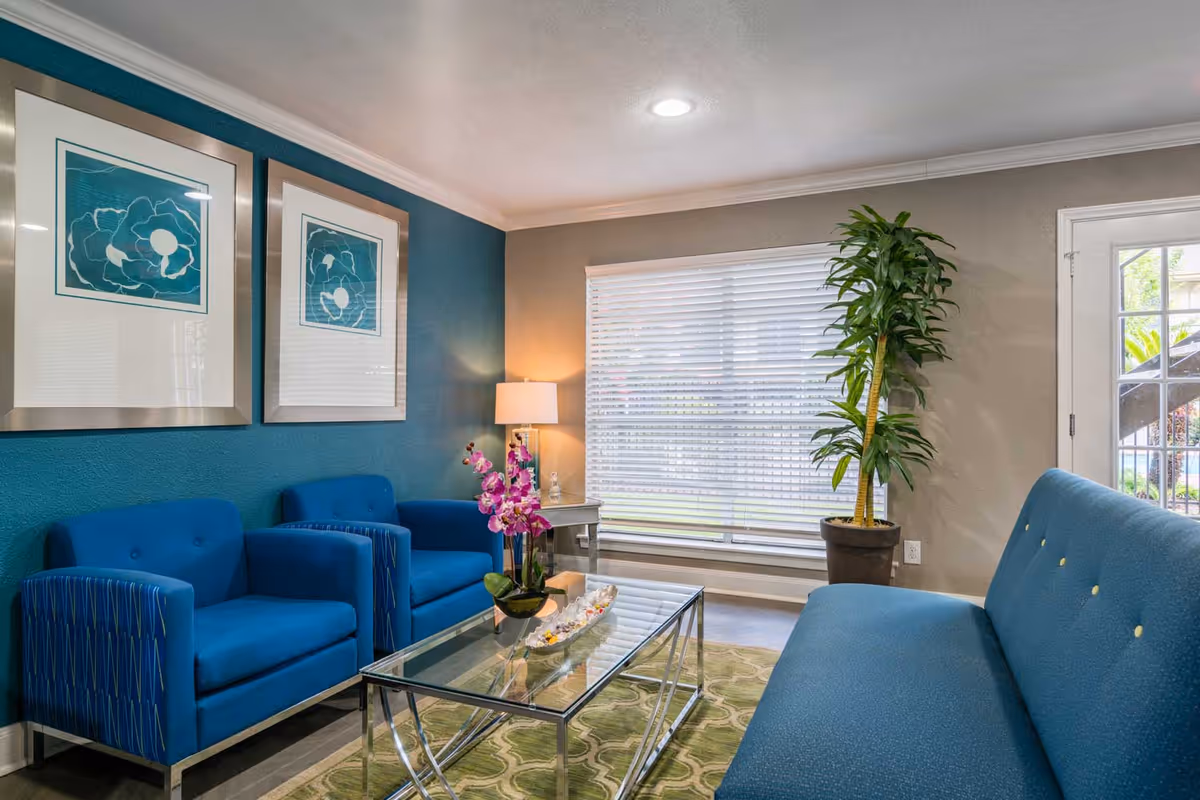 A bright and cozy living room with blue upholstered seating including two armchairs and a sofa. A glass coffee table with decorative items sits on a patterned green rug. The walls are painted teal and beige, adorned with two framed abstract artworks. A tall potted plant stands near a window with white blinds, and a side table with a lamp is placed between the armchairs.
