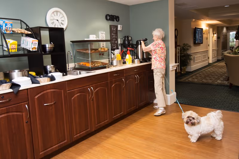 An elderly woman with white hair wearing a floral shirt and beige pants is pouring a drink from a coffee dispenser in a kitchenette area with wooden cabinets and a countertop. A small white dog on a blue leash stands on the wooden floor nearby. The kitchenette has a display case with pastries, a sink, plates, cereal boxes, and a clock on the wall. The background shows a hallway with carpet and wall decorations.