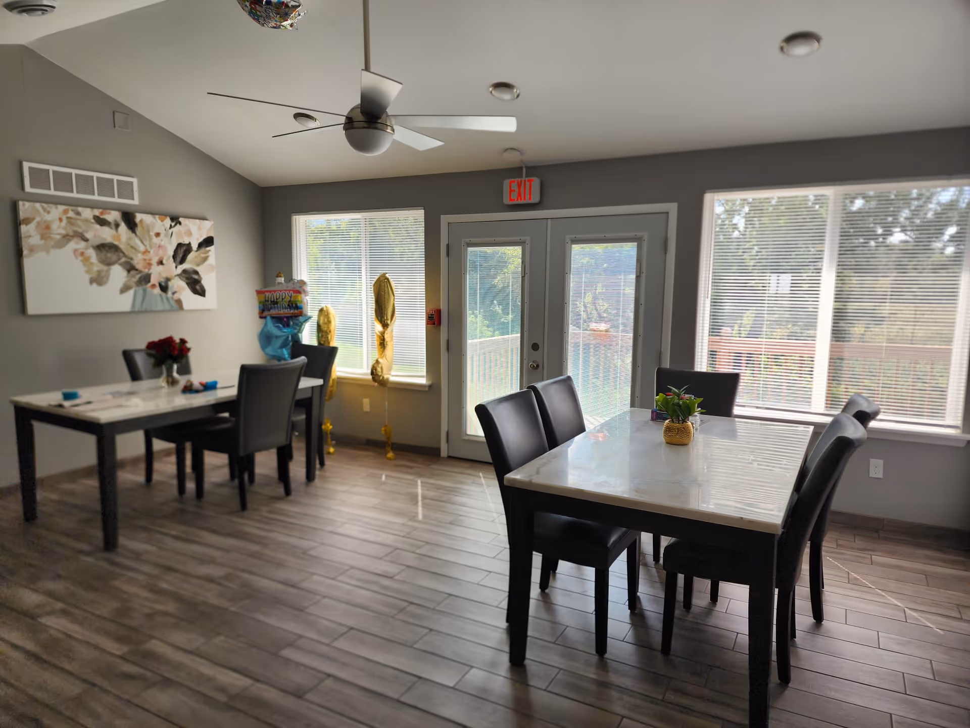 Dining area with two tables and several chairs, large windows and a glass door under a ceiling fan.