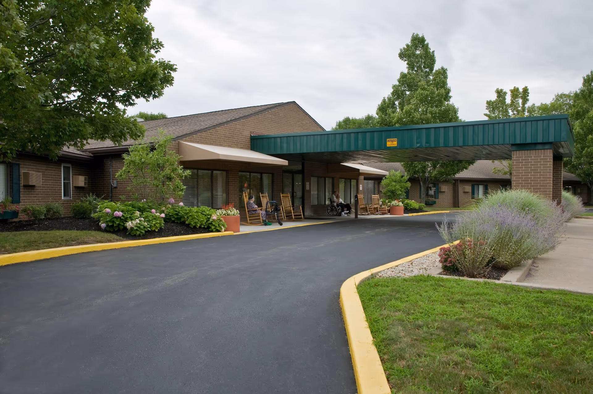 Covered entrance and driveway of a brick nursing care facility with rocking chairs, planters, and landscaped grounds.