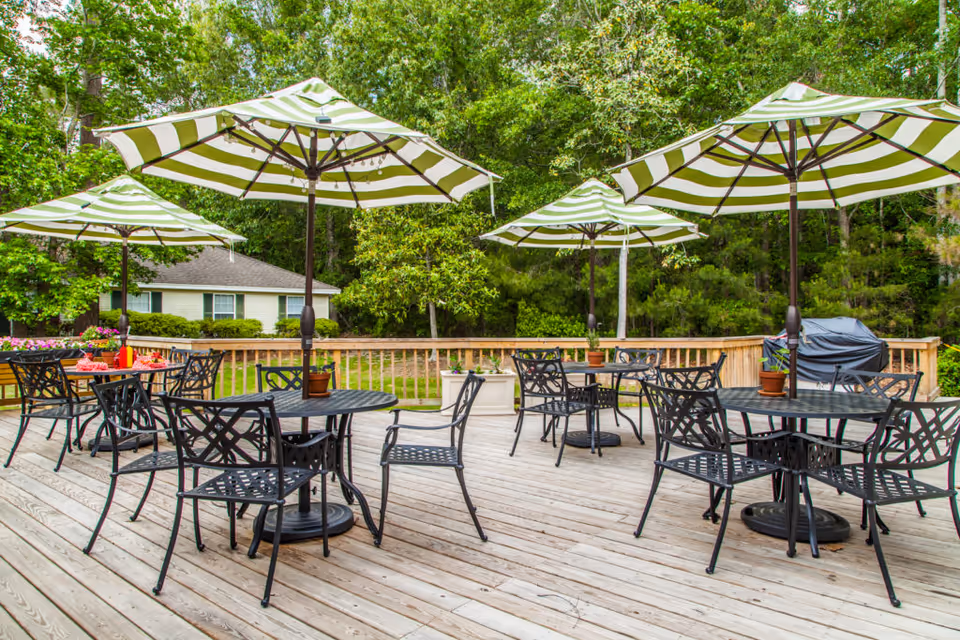 Outdoor wooden deck area with several black metal tables and chairs, each table shaded by a green and white striped umbrella. The deck is surrounded by a wooden railing and lush green trees and bushes are visible in the background. A covered grill is also seen on the right side of the deck.