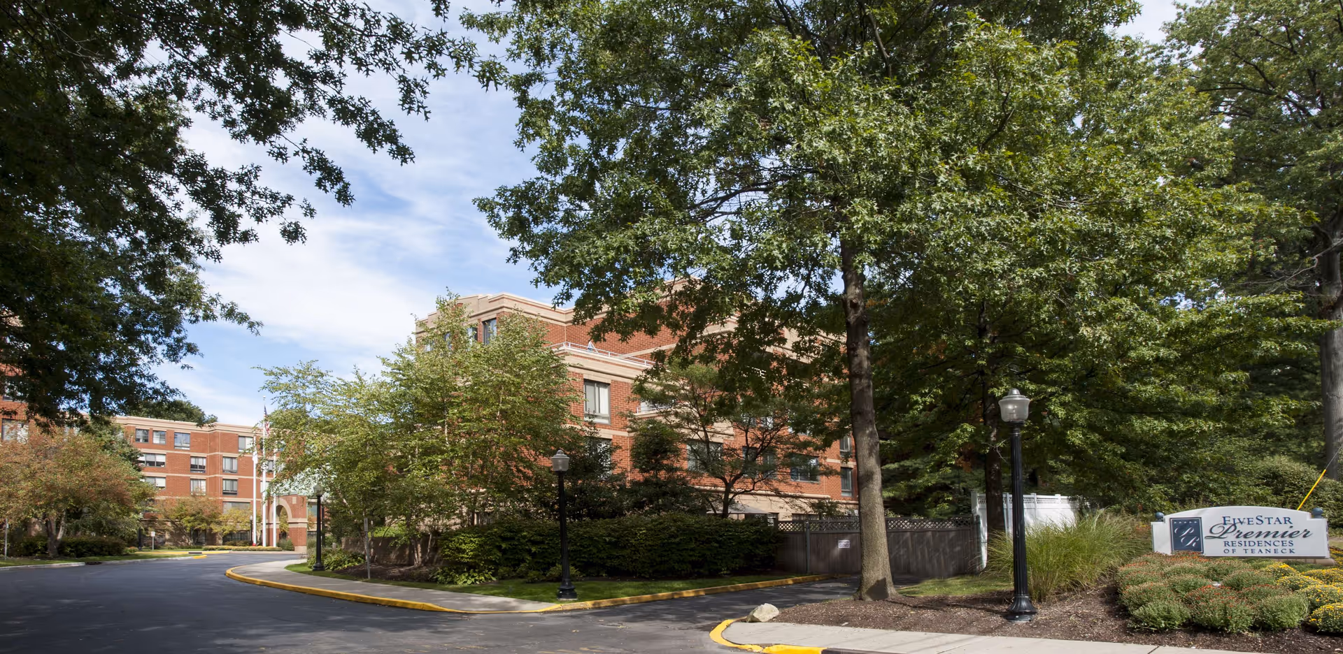 Exterior view of Five Star Premier Residences of Teaneck, showing a multi-story brick building surrounded by trees and landscaping, with a driveway and a sign displaying the facility's name.